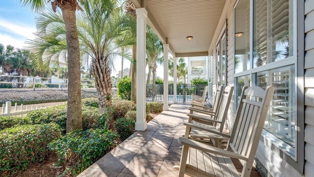 A covered outdoor area with wooden chairs, palm trees, and a view of a pool and buildings in the background. - Happy Hours (Destin)
