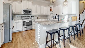 A modern kitchen with white cabinetry, a large island with bar stools, stainless steel appliances, and a hanging light fixture. - Happy Hours (Destin)