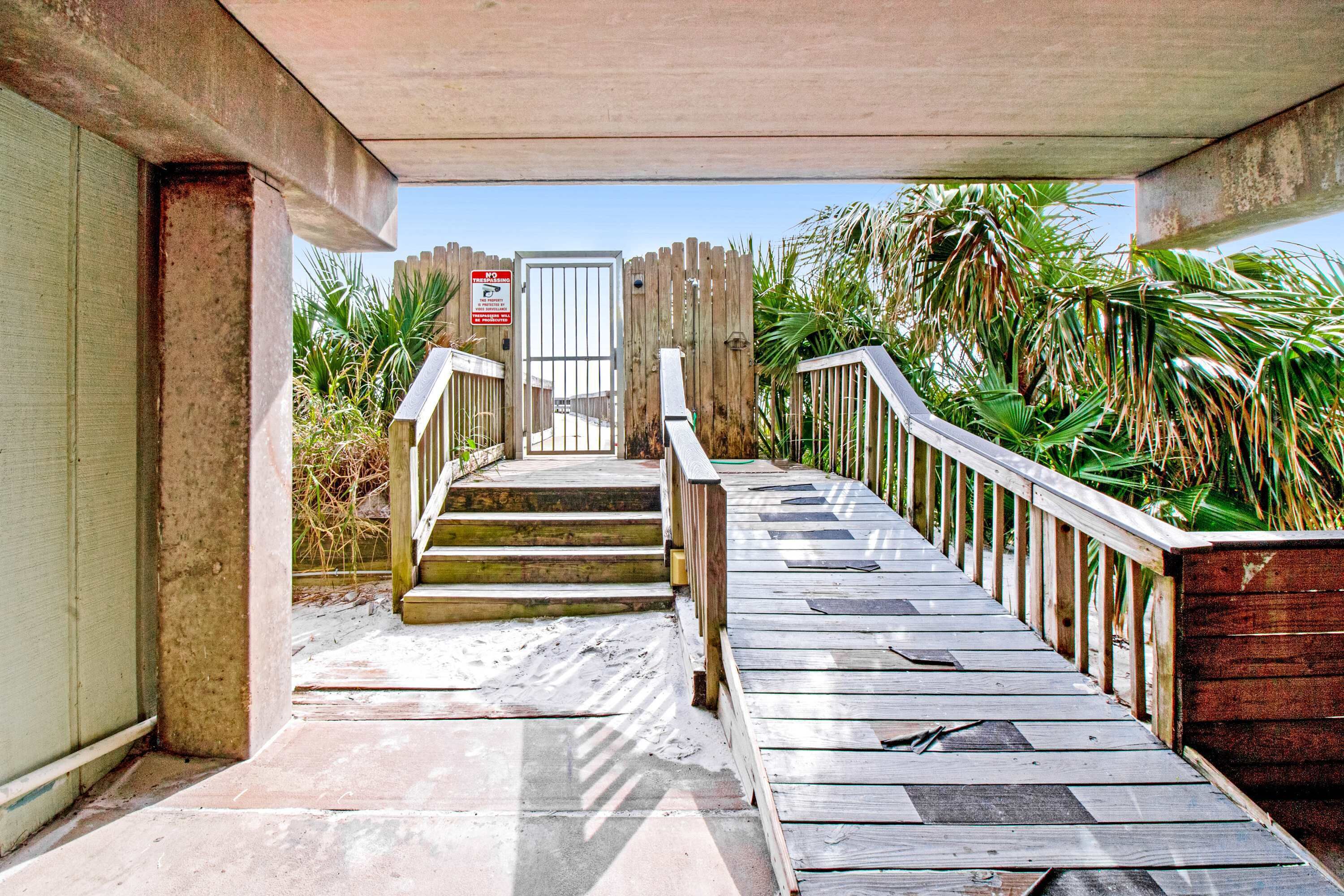 A wooden staircase leading to a gated entrance, surrounded by palm trees and a concrete structure.