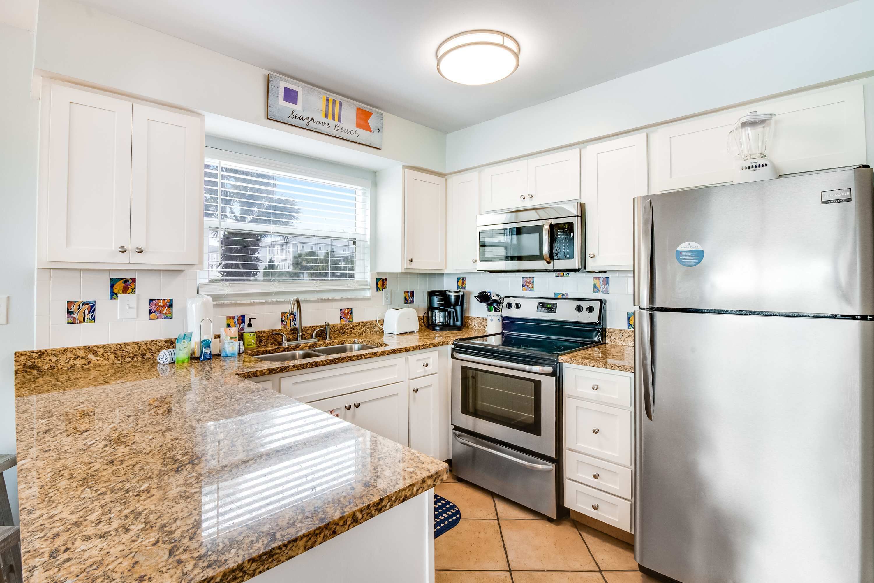 A modern kitchen with white cabinets, granite countertops, stainless steel appliances, and a window with blinds.