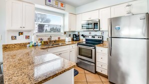 Ett modernt kök med vita skåp, bänkskivor i granit, rostfria vitvaror och ett fönster med persienner.