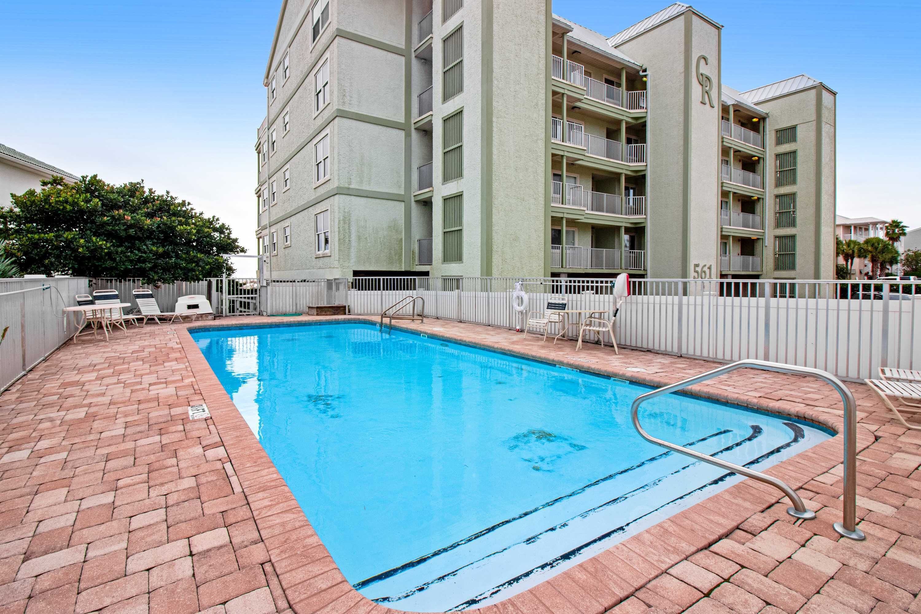 A swimming pool with a brick patio, surrounded by a white fence, and adjacent to a multi-story building with balconies.