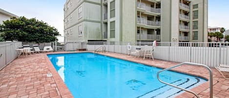 A swimming pool with a brick patio, surrounded by a white fence, and adjacent to a multi-story building with balconies.