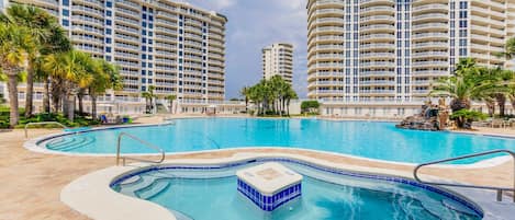 A large outdoor swimming pool surrounded by high-rise buildings, with palm trees and a clear sky.