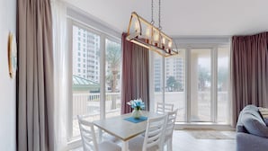 Uma sala de jantar com mesa e cadeiras, um lustre pendurado e grandes janelas com cortinas.