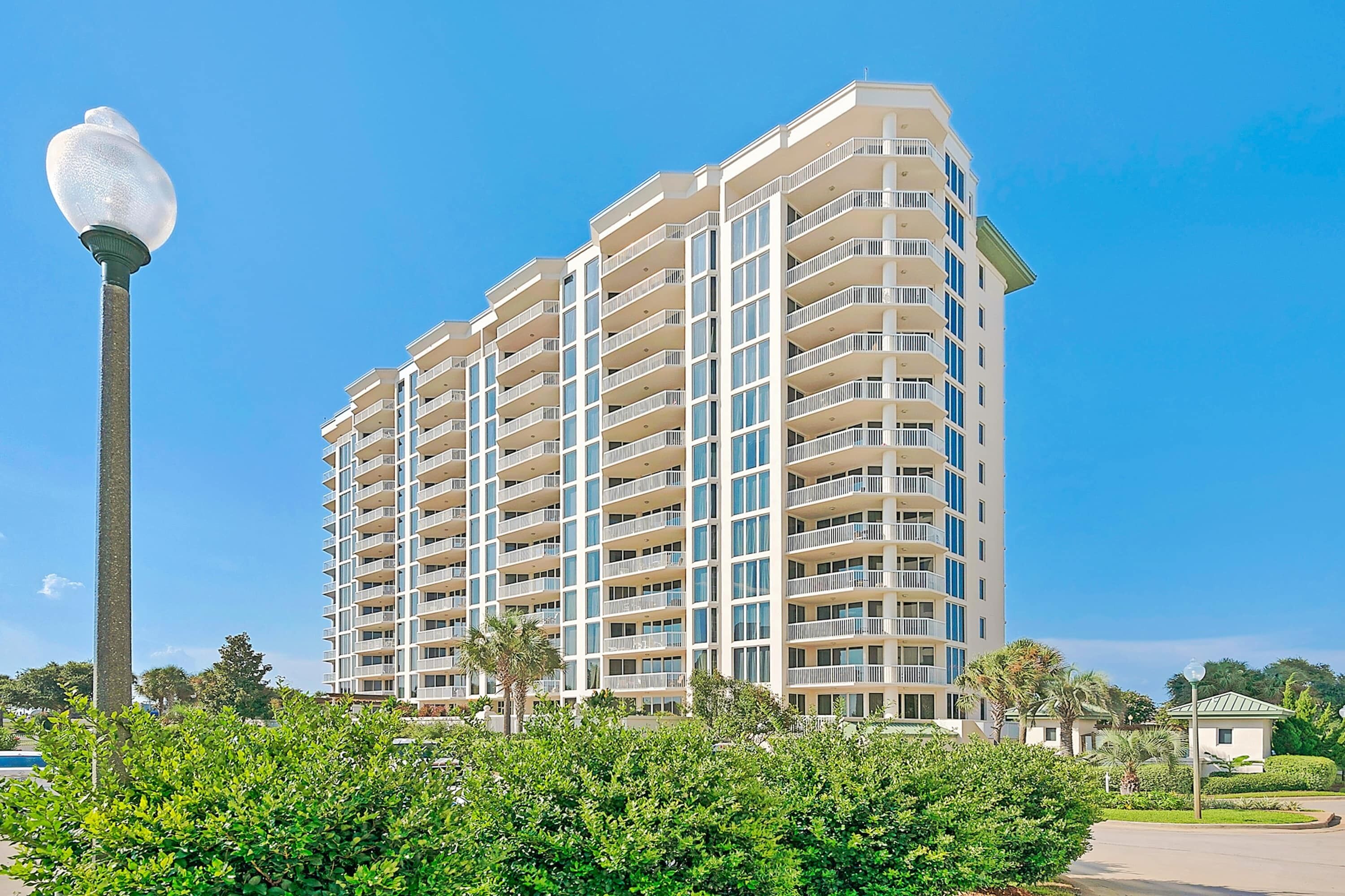 A multi-story hotel with a modern design, featuring balconies on each floor, surrounded by lush greenery.