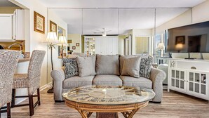 A living room with a round glass coffee table, a grey sofa, and a white cabinet with a television. - Sparkling Studio with Shared Pools, Hot Tubs, Golf, Tennis, & a Private Beach (Destin)