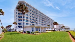 Un hotel de varios pisos con un diseño moderno, que cuenta con balcones, una entrada cubierta y un césped cuidado con palmeras.