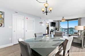 A modern living room with a glass-top dining table, comfortable seating, a chandelier, and a view of the outdoors. - 10th Floor Beautiful Condo, Splash pad w multiple pools & hot tub (Destin)