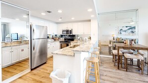 A modern kitchen with white cabinets, stainless steel appliances, a marble countertop, and a wooden dining area with stools. - Captiva beachfront condo w ocean views and pool & fitness center access (Destin)