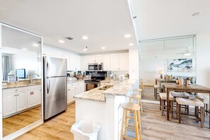 A modern kitchen with white cabinets, stainless steel appliances, a marble countertop, and a wooden dining area with stools. - Captiva beachfront condo w ocean views and pool & fitness center access (Destin)