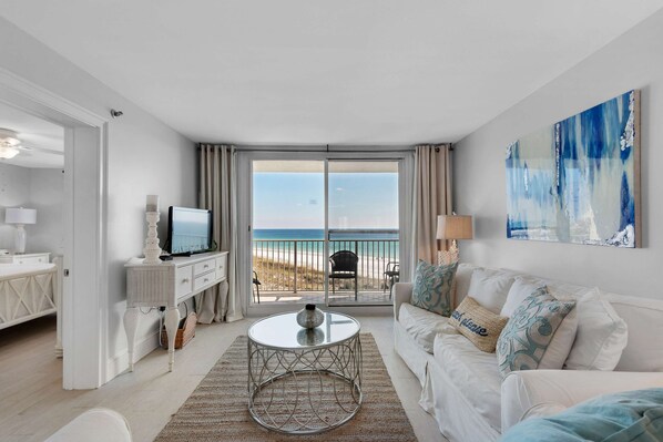 Sala de estar moderna com varanda vista para a praia, televisão e sofá branco com almofadas decorativas.