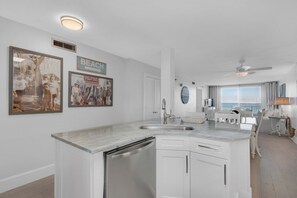 A modern kitchen with white cabinetry, a built-in dishwasher, and a large island with a sink. There are framed pictures on the wall, a ceiling fan, and a view of the ocean through large windows. - Amazing 5th Floor Condo Gulf Front, Pool, Beach Access, Near Shopping/Dining (Destin)