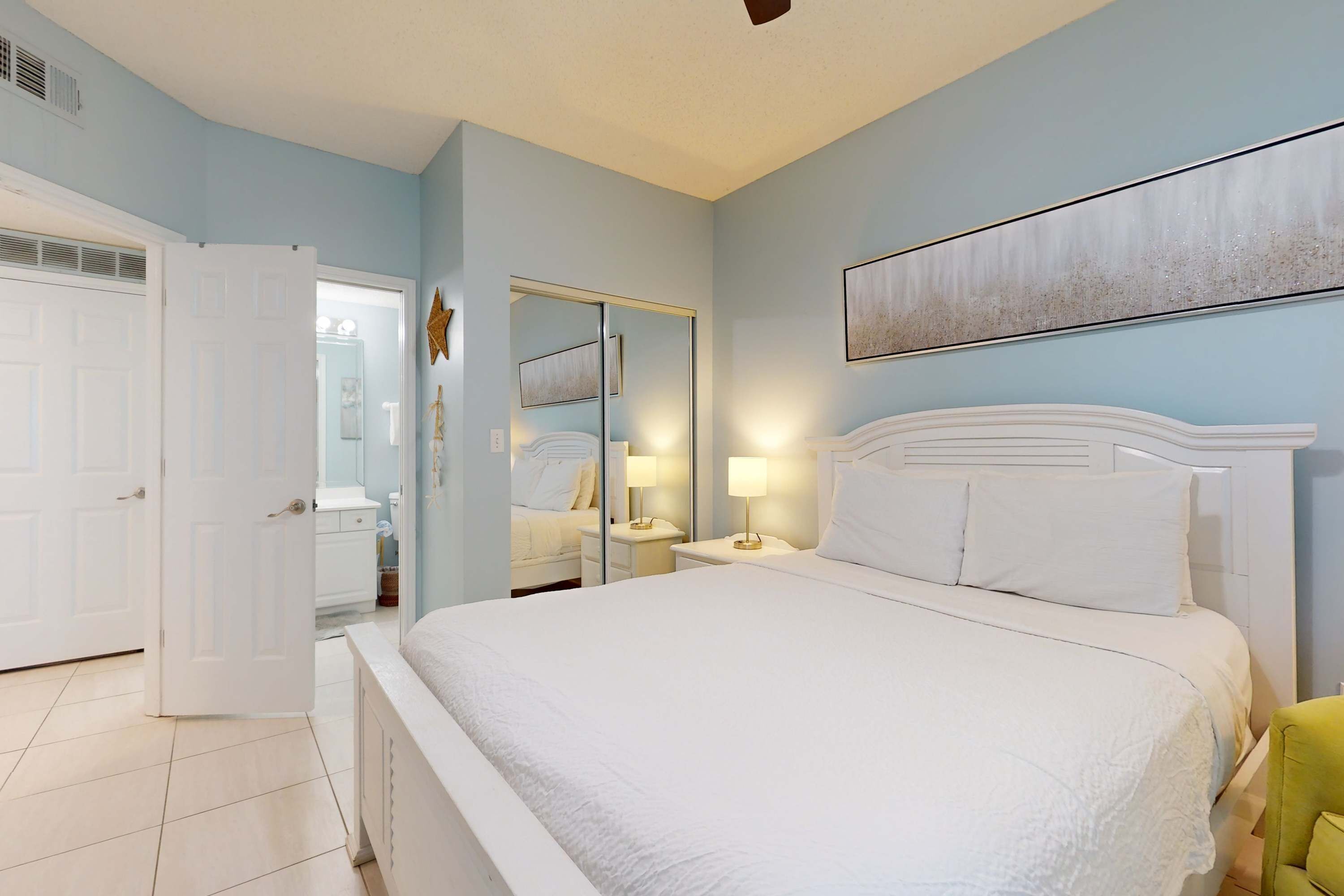 Une chambre à coucher avec un grand lit, une table de chevet avec une lampe, un grand miroir et une porte donnant sur une autre pièce.