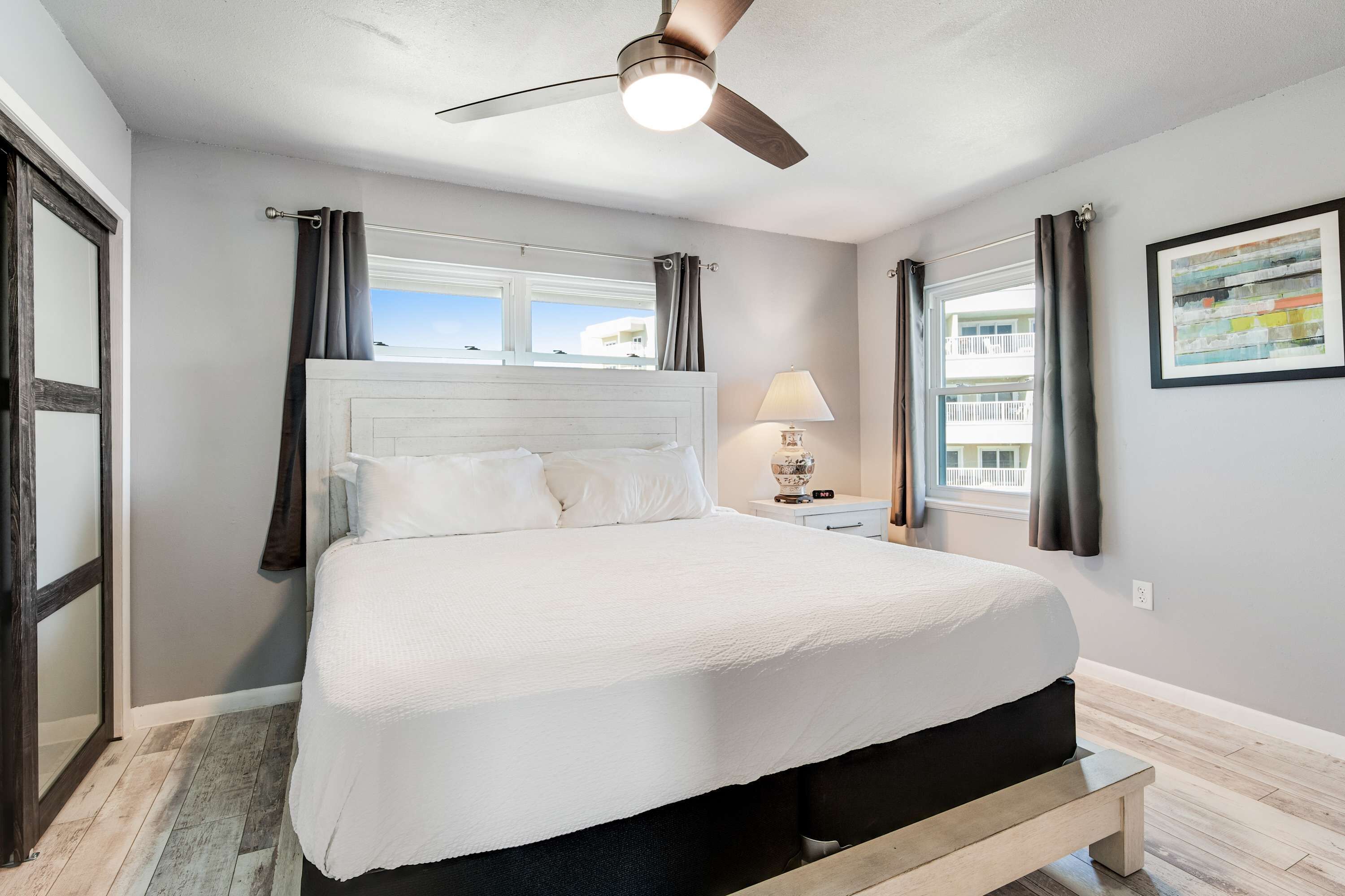 A bedroom with a bed, a ceiling fan, a nightstand with a lamp, and a window with curtains.