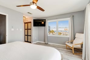 Une chambre à coucher comprenant un lit, un ventilateur de plafond, une télévision, une fenêtre avec vue et un fauteuil.