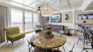Una habitación amplia con un gran ventanal, una mesa redonda de mármol con un centro de frutas, un sillón capitonado y una araña.