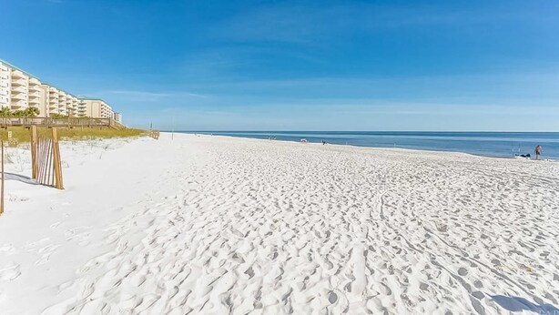 Une plage de sable avec des empreintes, un ciel bleu dégagé et, au loin, un bâtiment doté de balcons.