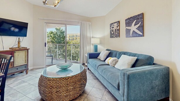 Un salón con sofá azul, una mesa de centro redonda de fibra natural, televisor de pantalla plana y balcón con vistas a edificios y árboles.