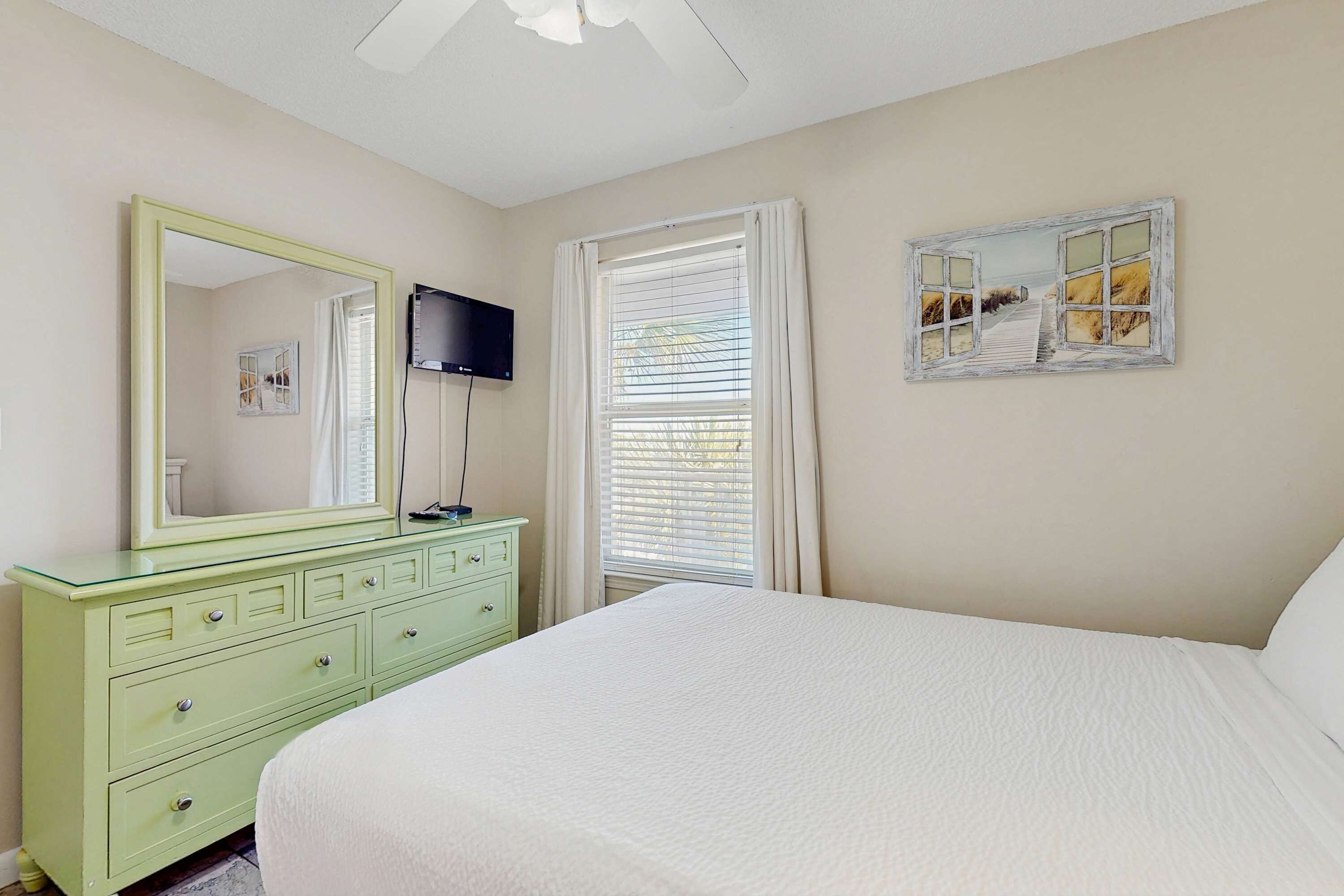 A bedroom with a bed, a dresser, a mirror, a television, and a window with curtains.