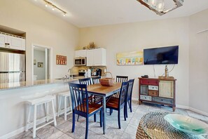 A modern kitchen with white cabinets, a wooden dining table, and blue chairs. A flat-screen TV is mounted on the wall above the table. - Bright, coastal condo with resort pool and hot tub (Miramar Beach)