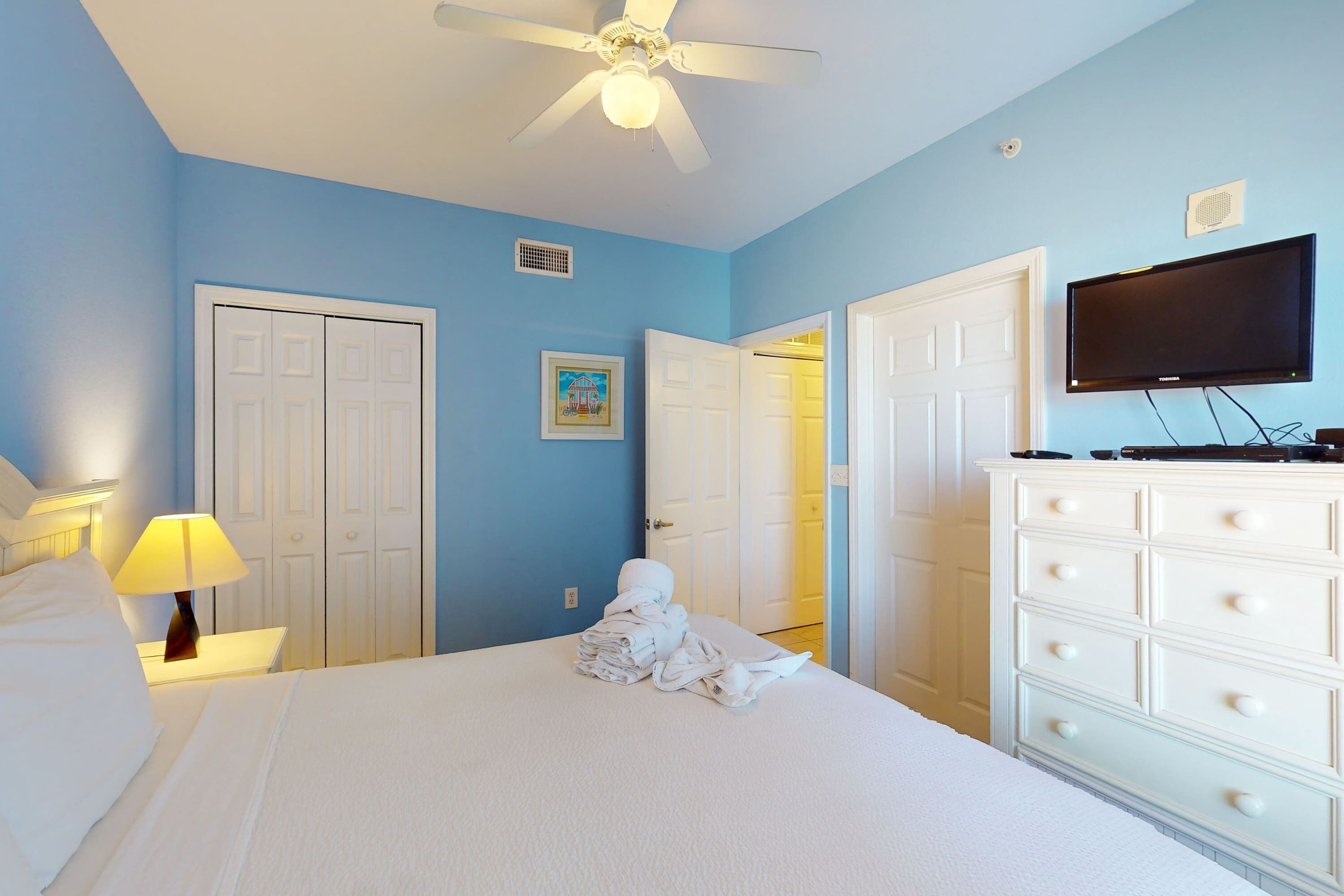 A hotel room with a bed, a ceiling fan, a television, and a dresser.