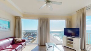 Ein geräumiges Wohnzimmer mit einem roten Ledersofa, einem Glastisch, einem Flachbildfernseher auf einer weißen Unterhaltungseinheit und einem Balkon mit Blick auf Strand und Meer.
