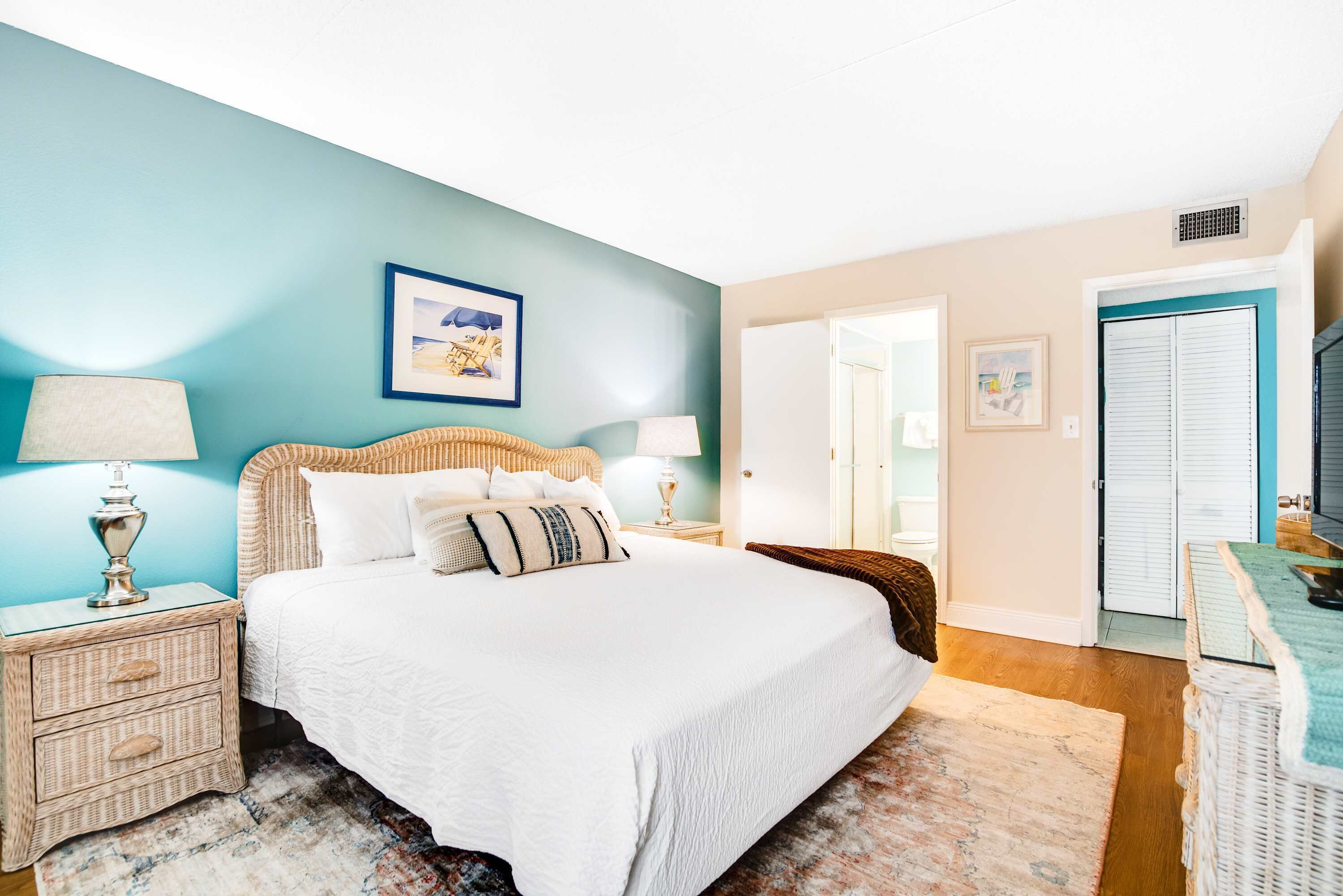 A bedroom with a bed, two nightstands, a lamp, a wicker chair, and a door leading to another room.