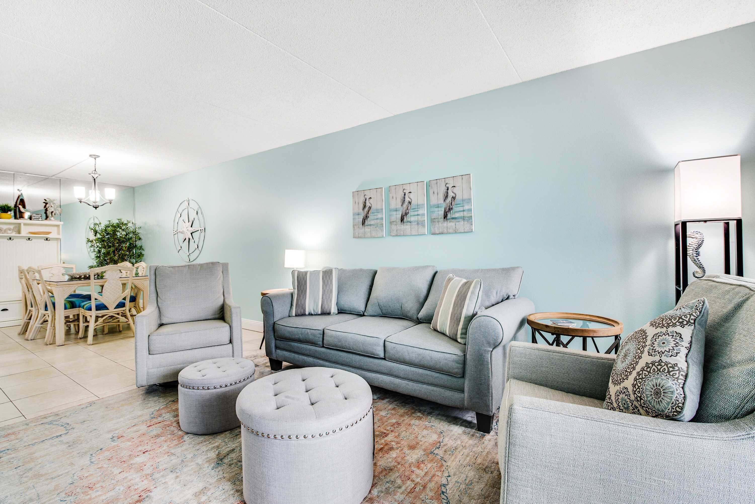 Ein Wohnzimmer mit einem grauen Sofa, zwei Sesseln, einem runden Hocker und einem Couchtisch. Über der Couch befinden sich Wandbilder und von der Decke hängt ein Kronleuchter.