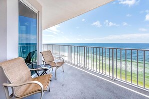 Een balkon met twee stoelen en een tafel, uitkijkend op een strand met de oceaan en een heldere lucht.