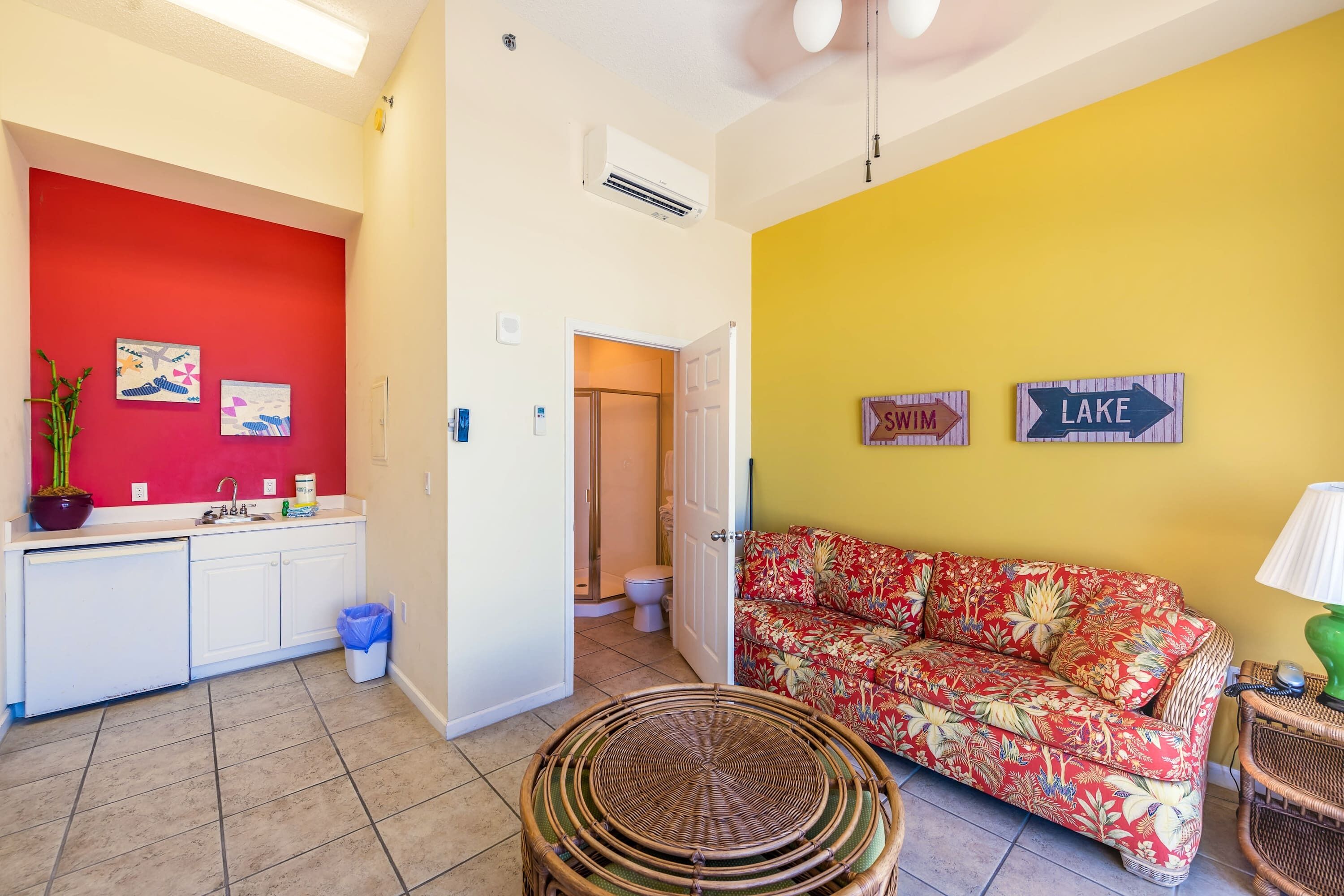 A living room with a red wall, a yellow wall, a floral sofa, a wicker coffee table, and a kitchen area with white cabinets.