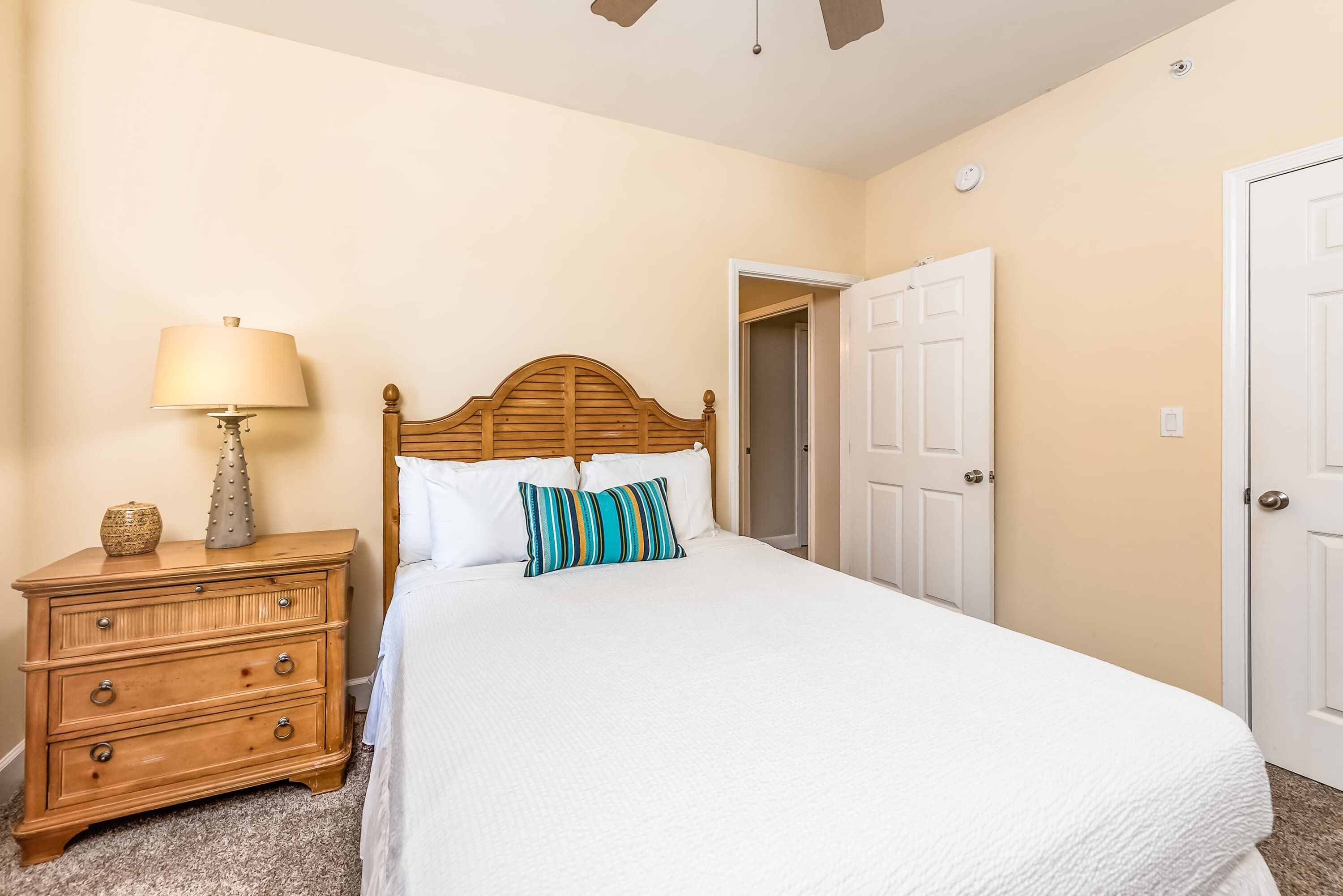 A bedroom with a wooden headboard bed, white bedding, a nightstand with a lamp, and a door.