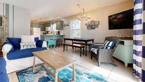 A living room with a sofa, coffee table, and a dining table with benches. There is a TV mounted on the wall and a window with blinds. - Beachside Villas | 1st Floor Condo | 6-min Walk to Beach | Pools (Santa Rosa Beach)