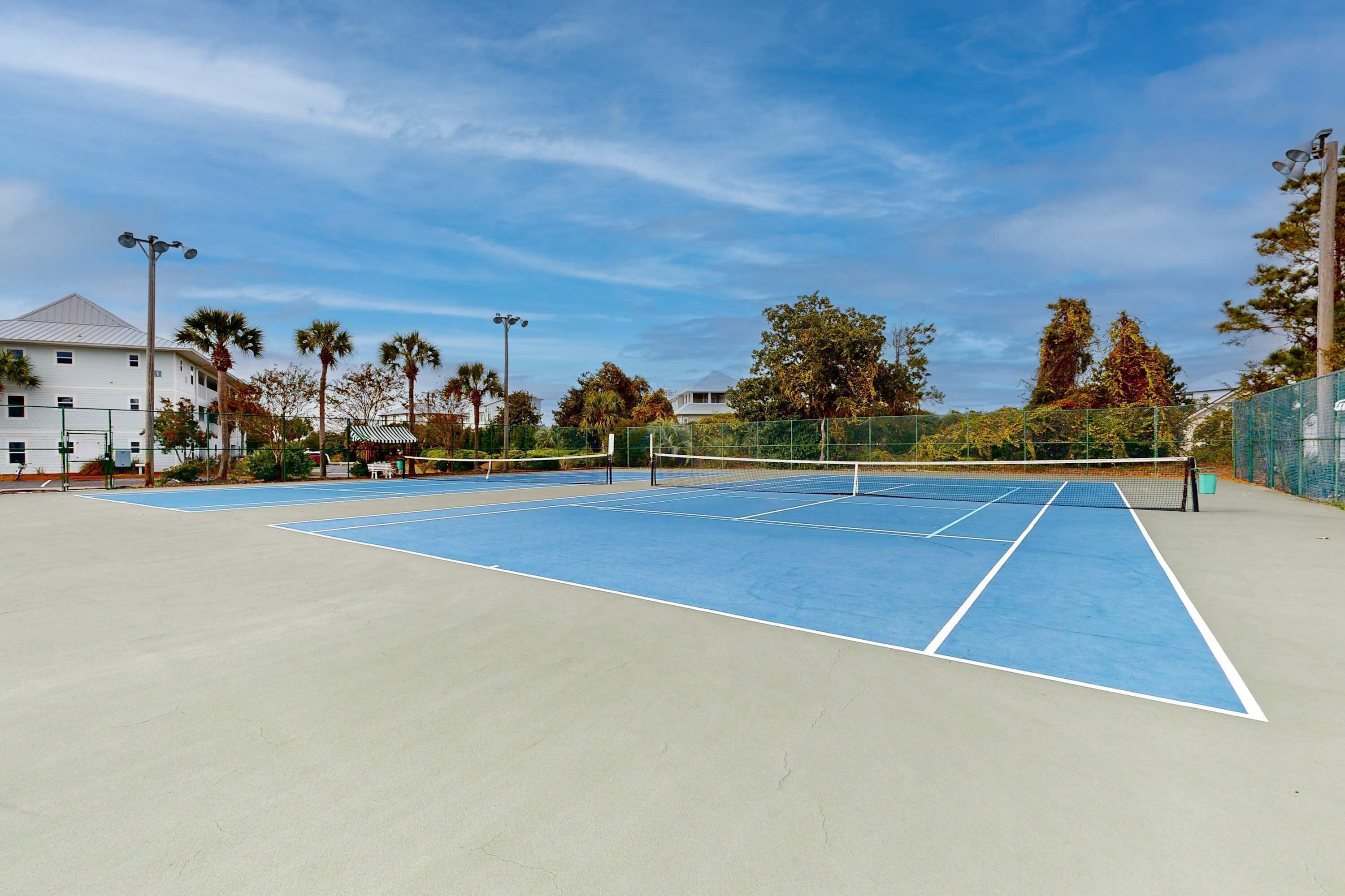 An outdoor tennis court with a net, surrounded by a fence and trees.