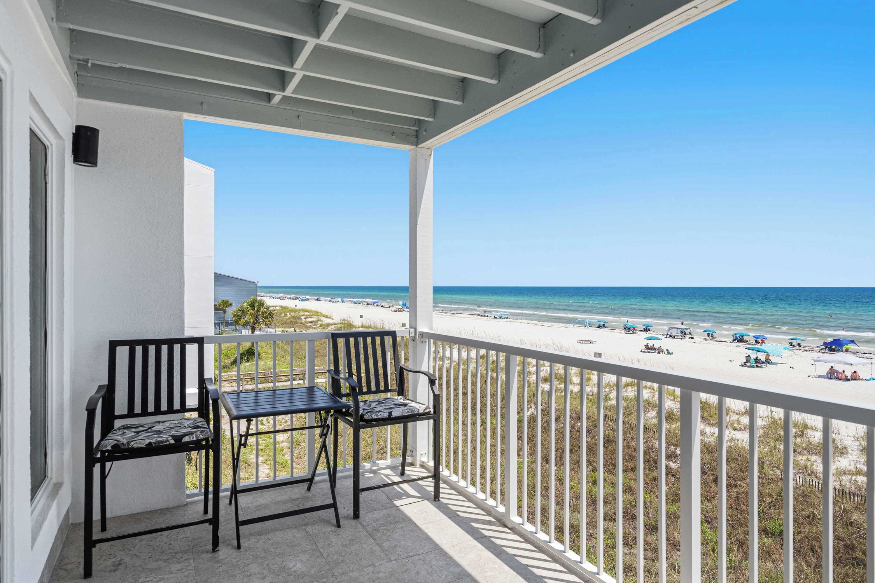 Ein Balkon mit zwei Stühlen und einem Tisch, mit Blick auf einen Strand mit Sonnenschirmen und Menschen.