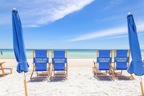 Vier blaue Strandstühle mit dem Aufdruck "Sterling Shores", auf einem sandigen Strand, im Hintergrund ein klarer, blauer Himmel.