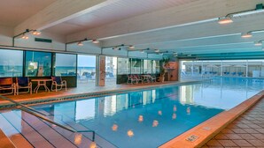 En indendørs swimmingpool med krystalklart vand, omgivet af liggestole og borde, samt udsigt til byen gennem store vinduer.