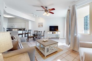 Ein modernes Wohnzimmer mit Sofa, Couchtisch, Esstisch, Stühlen und einem Fernseher. Es gibt einen Deckenventilator, eine Küchenzeile und ein Fenster mit Vorhängen.