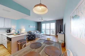 A modern kitchen with a central island, a dining area with a wooden table and chairs, a sofa, and a large window with a view. - Deluxe condo with Gulf view, heated pool & balcony (Panama City Beach)