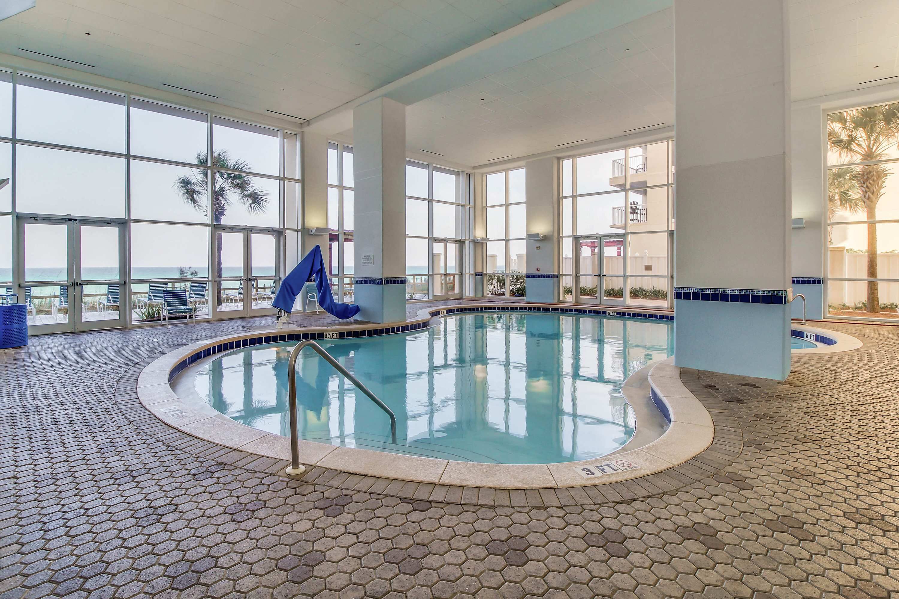 Une piscine intérieure offrant une vue dégagée sur l’océan grâce à de grandes fenêtres, un toboggan bleu et un sol carrelé.