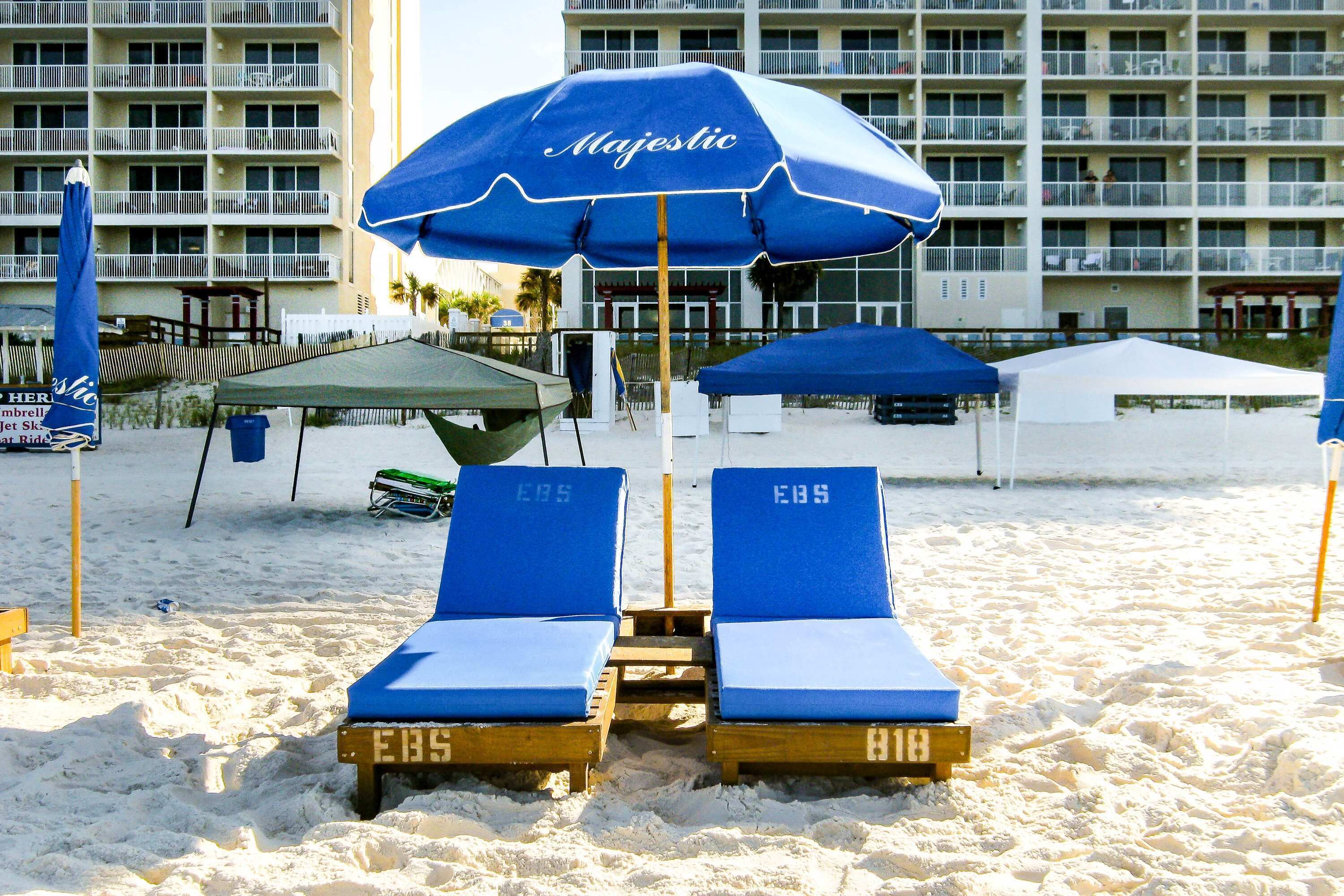 Een strand met twee blauwe ligstoelen met 'EBS' erop, onder een blauwe parasol waarop 'Majestic' staat. Op de achtergrond staat een gebouw met balkons.