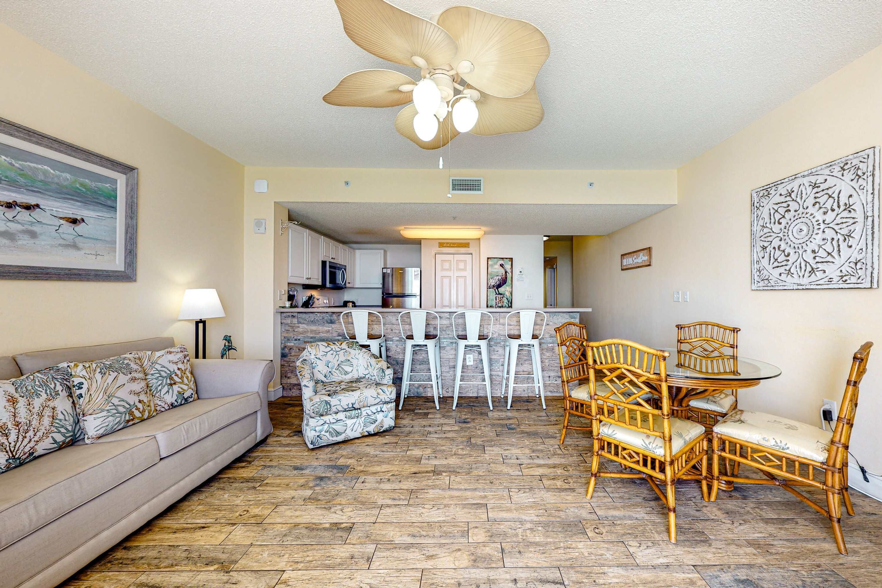 A living room with a sofa, a patterned armchair, a dining table with chairs, and a ceiling fan.