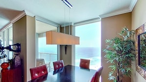 A dining area with a large table, red upholstered chairs, a potted plant, and a view of the ocean through large windows. - Stunning corner-condo with panoramic views, wraparound deck & beach access (Panama City Beach)