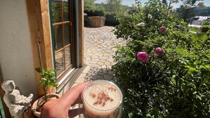 Une main tenant un verre contenant une boisson, une terrasse dallée de pierres, un bâtiment en bois et un jardin parsemé de fleurs roses.