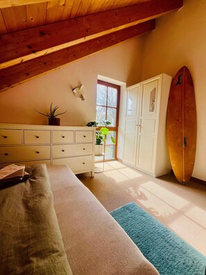 Une chambre avec un grand lit, une commode, une planche de surf et une fenêtre donnant sur un beau paysage.