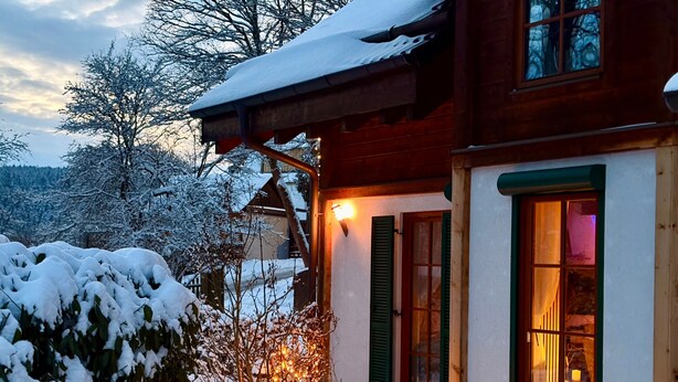 Une maison en bois, dont le toit est recouvert de neige et l’entrée est illuminée.