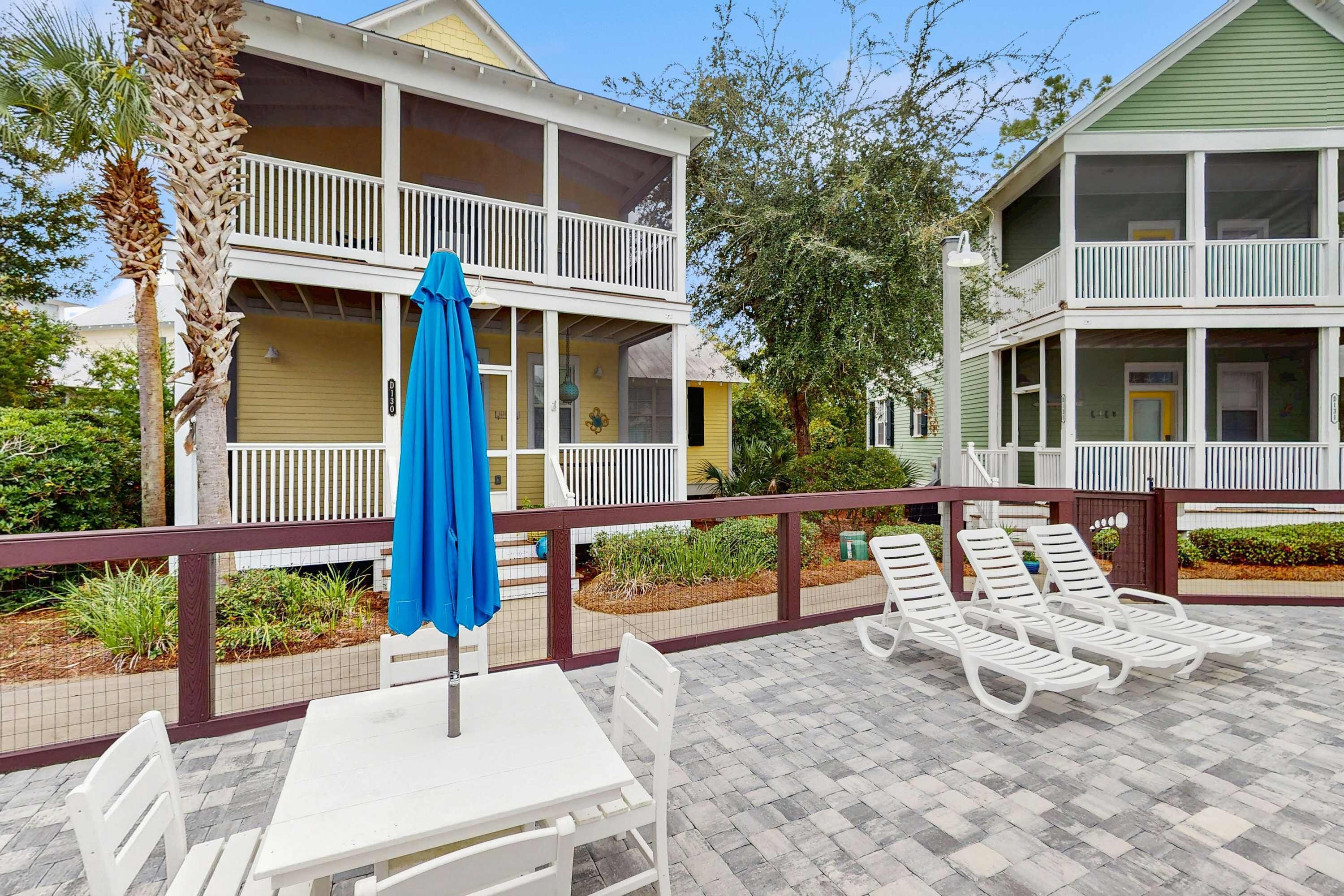 A hotel with a two-story balcony, outdoor seating area with white chairs and a blue umbrella, and a well-maintained garden.