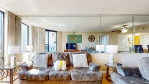 Ein modernes Wohnzimmer mit einer großen Couch, einem kleinen Sessel, einem Couchtisch und einem Fernseher. Eine Glaswand mit Spiegelung des Raumes ist vorhanden.