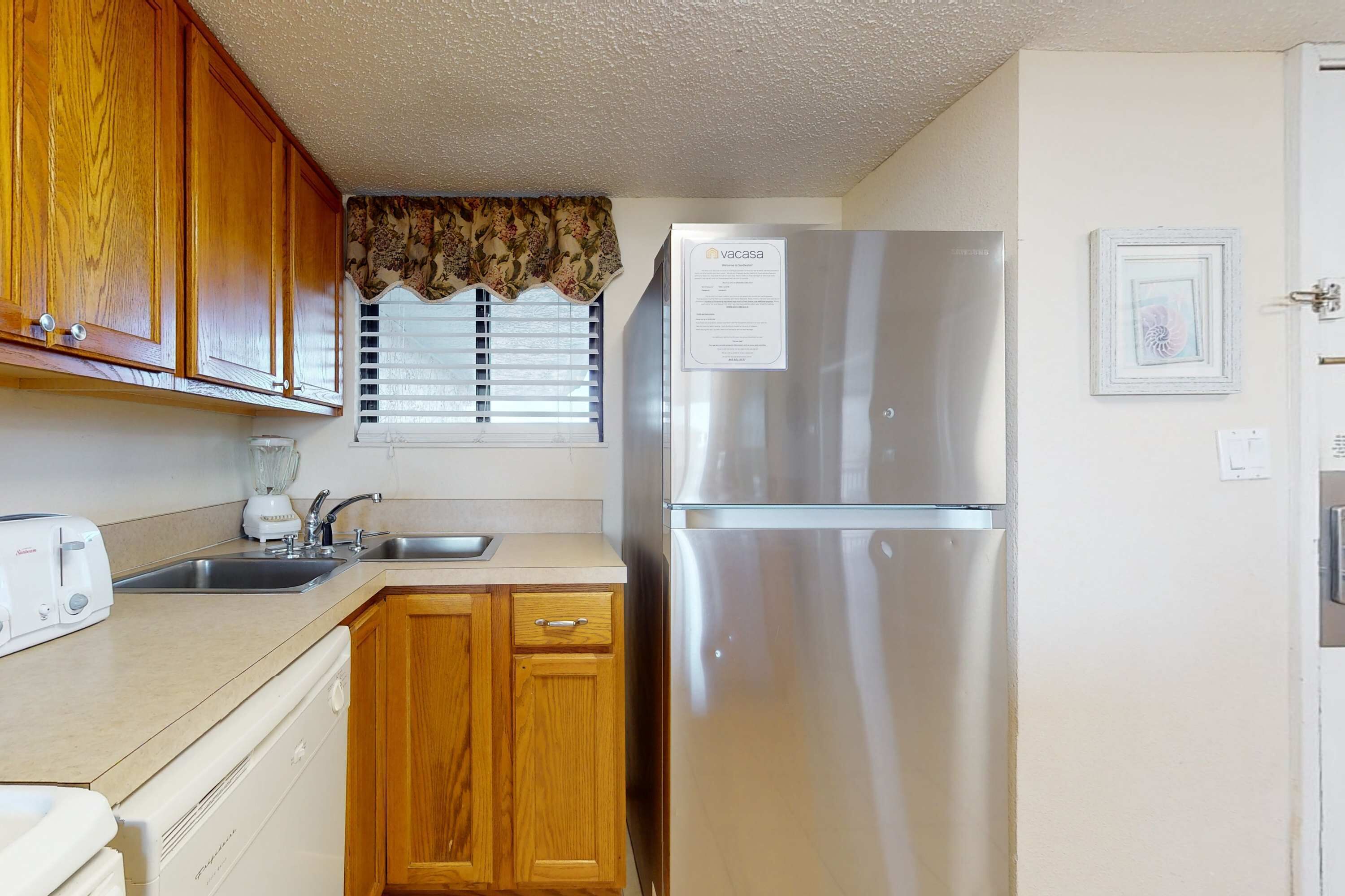 A compact kitchen with wooden cabinets, a stainless steel refrigerator, a built-in dishwasher, and a window with blinds.