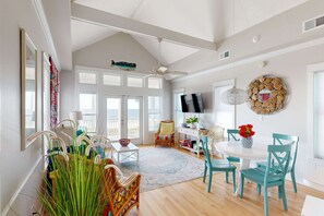 A spacious room with a high ceiling, large windows, a ceiling fan, and a dining area with teal chairs and a white table. - Beachfront home with expansive deck, screened porch, washer&dryer & central AC (St. George Island)